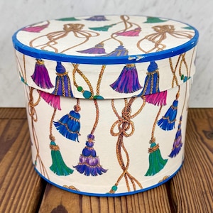 May include: A round hat box with a blue rim and a repeating pattern of colorful tassels and rope. The tassels are in shades of blue, purple, pink, and green. The box is on a wooden surface.
