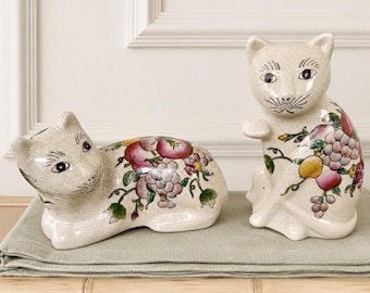 May include: Two ceramic cat figurines with a cream-colored crackle glaze finish. One cat is in a reclining position, while the other sits upright. Both feature hand-painted colorful illustrations of grapes, peaches, and green leaves on their bodies.