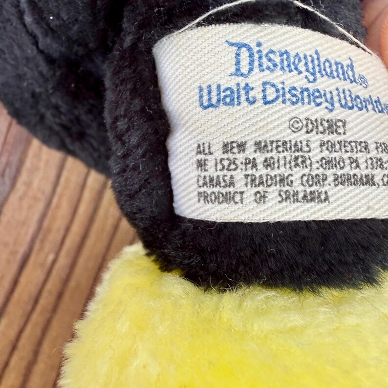 May include: Close-up of a plush toy tag. The tag is white with blue text that reads "Disneyland Walt Disney World" and "&copy;DISNEY". The tag also includes text about materials and the product's origin in Sri Lanka.