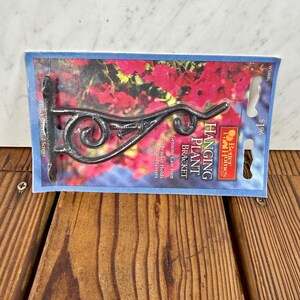 Könnte beinhalten: Ein schwarzer, dekorativer Hängepflanzenhalter aus echtem Gusseisen. Die Halterung hat ein gerolltes Design und ist auf einer blau-roten Karte verpackt. Auf der Karte steht "HANGING PLANT BRACKET" und "Bracket Holds Small Planters."