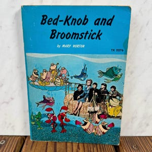 May include: Vintage paperback book titled "Bed-Knob and Broomstick" by Mary Norton. The cover features an illustration of a whimsical underwater scene with cartoon characters, fish, and people. The book has a blue background.