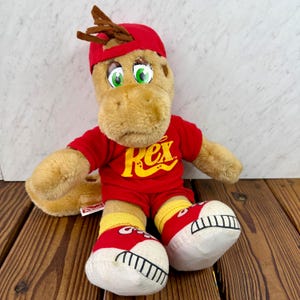 May include: A plush toy dinosaur wearing a red baseball cap, red shirt and shorts with the word "Rex" in yellow, and red sneakers with white soles. The dinosaur has green eyes and brown hair.