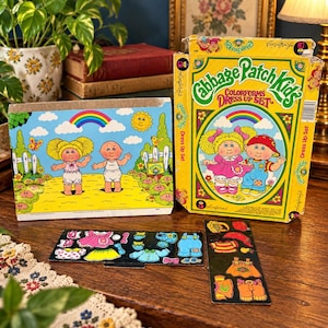 May include: A Cabbage Patch Kids Colorforms Dress Up Set featuring a yellow box with illustrations of two dolls, a play board showing a sunny outdoor scene with a rainbow, and two black sheets containing various colourful clothing stickers.