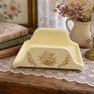 May include: A cream-colored ceramic napkin holder with a scalloped edge and a floral decal featuring a central pink flower surrounded by green leaves and smaller white blossoms. The holder has a smooth, glossy finish and a curved, elevated back.