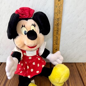 May include: A Minnie Mouse plush doll, featuring a red dress with white polka dots, a red bow, and black ears. The doll has white gloves and yellow shoes. A wooden ruler is in the background.