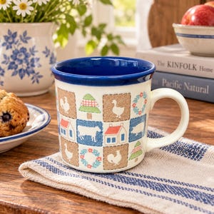 May include: A ceramic mug with a cream-colored base and a deep blue interior rim. The exterior features a repeating grid pattern of squares containing illustrations of farm animals, houses, pine trees, and floral wreaths in blue, red, and green.