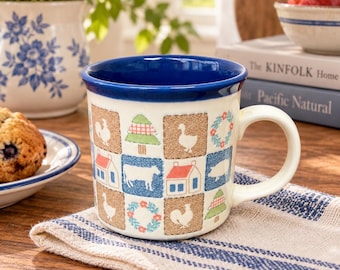 May include: A ceramic mug with a cream-coloured base and a deep blue interior rim. The exterior features a repeating grid pattern of squares containing illustrations of farm animals, houses, pine trees, and floral wreaths in blue, red, and green.