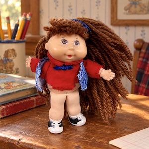 May include: A doll with a round face, brown yarn hair, and a blue hair ribbon. The doll wears a red long-sleeved shirt with a blue collar, a blue vest, beige pants, and black and white shoes. The doll has a soft body and plastic head.