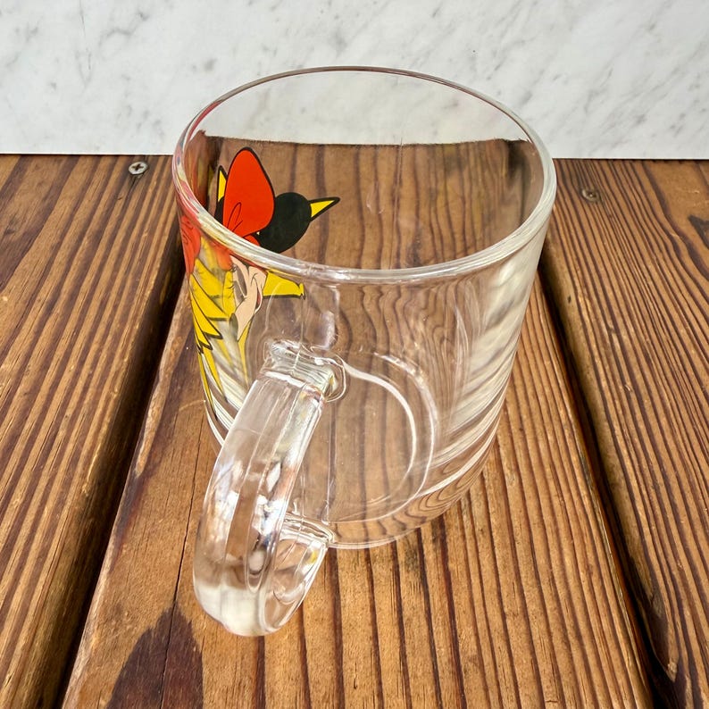May include: A clear glass mug featuring a colorful graphic of a stylized bird with a bright red crest, yellow feathers, and black accents. The mug has a smooth, transparent handle and a cylindrical body, showcasing the vibrant illustration on its side.