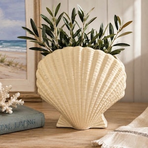 May include: A cream-colored ceramic vase shaped like a large scallop shell. The vase features a ribbed, fan-like texture and is filled with green olive branches. The shell-shaped vessel has a wide, curved top edge and a stable, flat base.