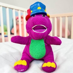 May include: A plush purple dinosaur toy with a green belly, yellow feet, and a blue police hat. The toy has an open mouth and outstretched arms, sitting on a white surface. The background includes a white crib.