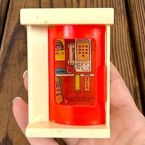 May include: A vintage Fisher-Price toy telephone booth. The toy has a cream-colored frame and a red interior with a graphic of a telephone, a woman, and the words "BIG CITY" and "ILLAGE". The toy is marked "997" and "73 FISHER-PRICE TOYS, EAST AURORA, N.Y."