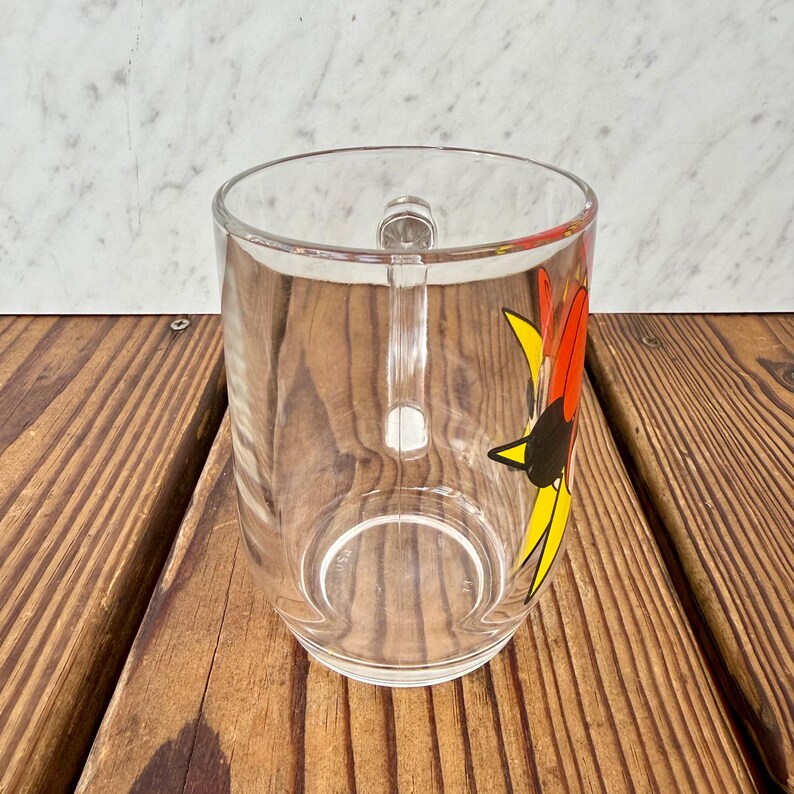 May include: A clear glass mug featuring a vibrant graphic design on its side. The illustration displays a stylized red, yellow, and black sun-like shape with sharp, pointed rays. The mug has a sturdy, integrated glass handle and a smooth rim.