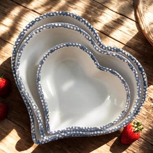 May include: Set of three heart-shaped ceramic bowls. The bowls are white with a blue dotted trim. The bowls are nested on a wooden surface, with a few strawberries scattered around.