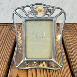 Op de afbeelding: Een fotolijst in vintage-stijl met een zilverkleurig metalen frame en helder glas. Het glas is versierd met gedroogde bloemen en bladeren. De lijst heeft een rechthoekige opening voor een foto.