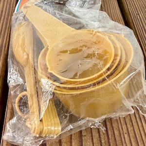 Può includere: Un set di misurini e cucchiai marrone chiaro, ancora nella loro confezione di plastica originale. I misurini sono impilati e i cucchiai sono visibili a sinistra. Il set include varie misure per la cottura.