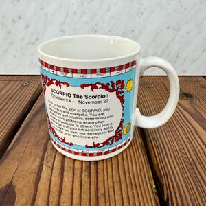 May include: A white ceramic mug featuring a light blue panel with red decorative scrollwork and the text SCORPIO The Scorpion October 24 - November 22. The design includes a border of red and white squares and astrological symbols.