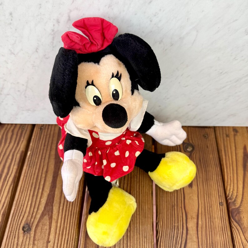 May include: A plush Minnie Mouse doll with a red bow, black ears, and a red polka dot dress. The doll has yellow shoes and white gloves. The doll is sitting on a wooden surface.