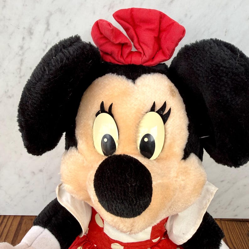 May include: A plush Minnie Mouse doll with black ears, a red bow, and a red and white polka dot dress. The doll has large, expressive eyes and a black nose. The doll is a classic Disney character.
