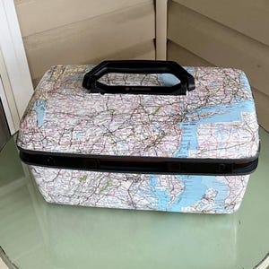 May include: A vintage Samsonite suitcase with a map design. The suitcase has a black handle and trim, and the map covers the entire surface. The map includes the New York area. The suitcase is sitting on a glass table.