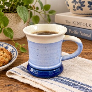 Könnte beinhalten: Ein Keramikbecher mit dreifarbigem Design, bestehend aus einem weißen Rand, einem hellblauen, gesprenkelten Mittelteil und einem dunkelblauen Boden. Der Becher hat einen geschwungenen Henkel, enthält eine dunkle Flüssigkeit und steht auf einem beigen Tuch.