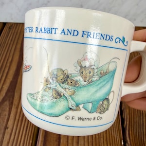 May include: White ceramic mug featuring the text "PETER RABBIT AND FRIENDS" in blue. The mug has a blue border and a whimsical illustration of mice in a blue shoe. The mug is marked with "© F. Warne & Co."