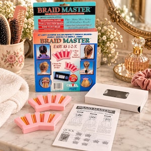 May include: The Braid Master hair braiding system includes a cardboard box with product text, two pink and orange plastic hair styling tools, a white instructional booklet, and a white box containing a VHS tape for hair styling guidance.
