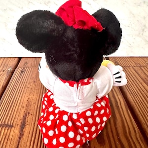 K&ouml;nnte beinhalten: Eine Minnie Mouse Pl&uuml;schpuppe, von hinten gesehen, mit schwarzen Fellohren und Kopf, einer roten Schleife und einem wei&szlig;en Kleid mit rotem Tupfenrock. Die Puppe hat wei&szlig;e Handschuhe mit schwarzen Linien, vor einem Holz Hintergrund.