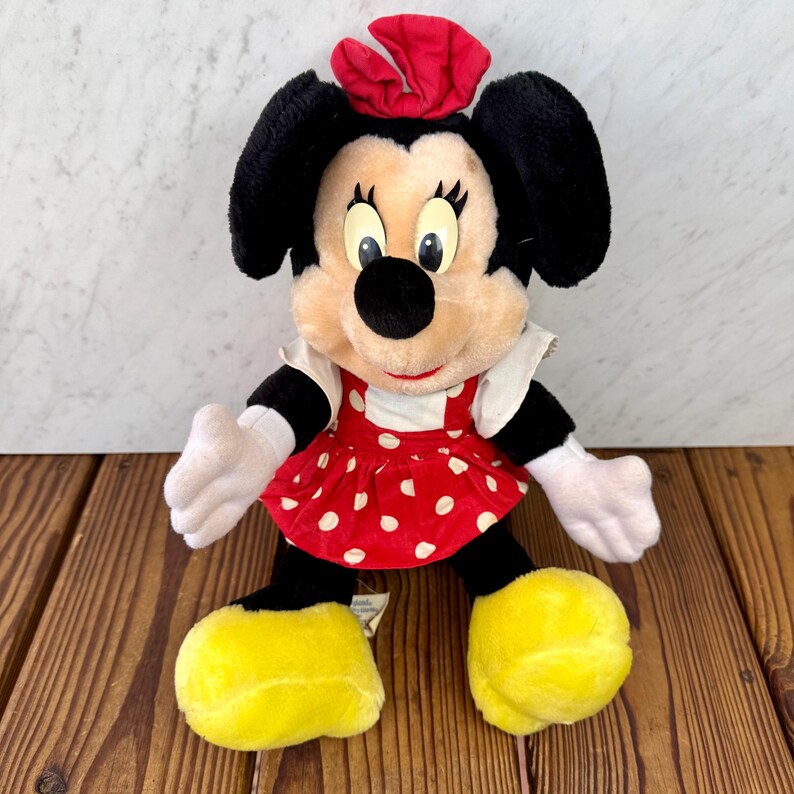 May include: A plush Minnie Mouse doll with black ears, a red bow, and a red dress with white polka dots. The doll has white gloves, black legs, and yellow shoes. The doll is sitting on a wooden surface.