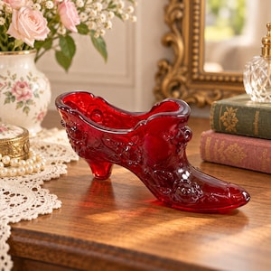 May include: A deep red glass shoe figurine featuring an intricate embossed floral pattern across its surface. The decorative object is shaped like a high-heeled pump with a pointed toe and a wide opening, showcasing a translucent finish and glossy texture.