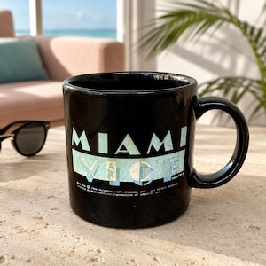 May include: A black ceramic mug featuring the text MIAMI VICE in a light blue, distressed font. The design includes a rectangular light blue background behind the word VICE. Small copyright text is printed at the bottom of the mug graphic.