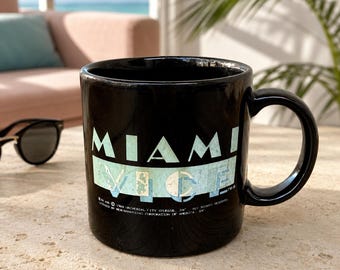 May include: A black ceramic mug featuring the text MIAMI VICE in a light blue, distressed font. The design includes a rectangular light blue background behind the word VICE. Small copyright text is printed at the bottom of the mug graphic.
