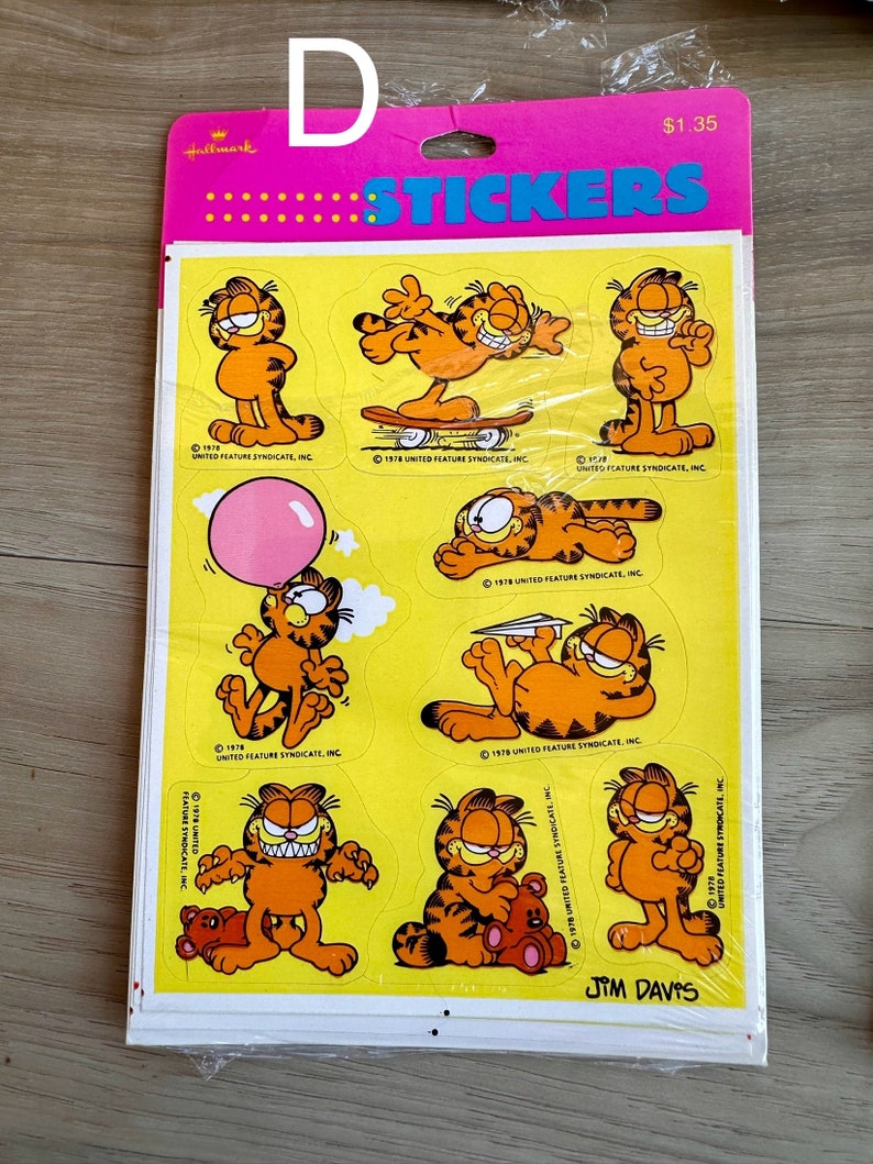 K&ouml;nnte beinhalten: Eine Bogen Garfield-Aufkleber mit dem orangefarbenen Tabby-Kater in verschiedenen Posen. Die Aufkleber befinden sich auf einem gelben Hintergrund mit dem Text "Stickers" und "Jim Davis", der auf dem Bogen gedruckt ist.