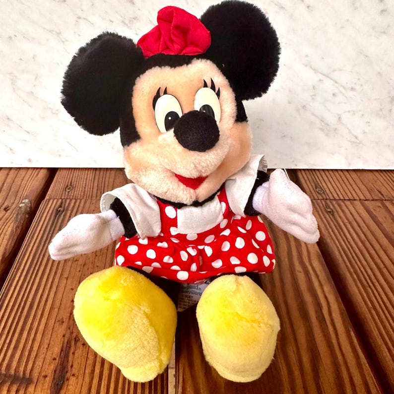 K&ouml;nnte beinhalten: Eine Pl&uuml;schpuppe von Minnie Mouse mit schwarzen Ohren, einer roten Schleife und einem rot-wei&szlig; gepunkteten Kleid. Die Puppe hat wei&szlig;e Handschuhe und gelbe Schuhe. Die Puppe sitzt auf einer Holzoberfl&auml;che.