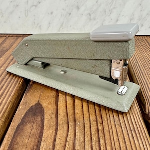 May include: A vintage, olive-green stapler with a light gray top. The stapler is made of metal and has a silver staple exit. The stapler is resting on a wooden surface.
