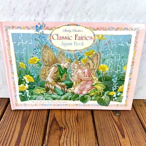 Puede incluir: Una caja rosa con un libro de rompecabezas titulado "Classic Fairies" con un borde floral. La portada representa a dos hadas con alas de mariposa en un jardín. La caja dice "With Four 96-Piece Jigsaws."