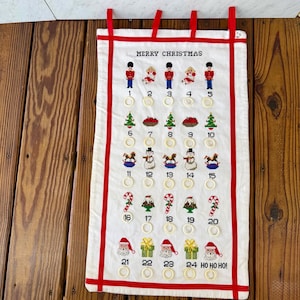 May include: A white fabric Christmas advent calendar with red trim and hanging loops. The calendar features embroidered images of soldiers, trees, snowmen, and Santa Claus, with numbers 1-24 and the text "MERRY CHRISTMAS" and "HO HO HO!"