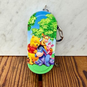 Può includere: Un portachiavi colorato con Winnie the Pooh e i suoi amici. Il design include Winnie the Pooh, Tigro, Pimpi ed Eeyore su uno sfondo di cielo blu e alberi verdi. Il portachiavi ha un anello in metallo color argento.