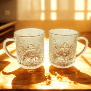 May include: Two clear glass mugs with textured surfaces and handles. Each mug features a detailed embossed design of a smiling face surrounded by floral elements. The mugs are set on a wooden surface.