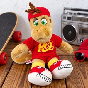 May include: A plush dinosaur toy wearing a red baseball cap, a red t-shirt with the yellow text Rex, and red and white sneakers. The toy has tan fur, large green eyes, and brown hair tufts. A small white tag with the text Rex is visible on the side.