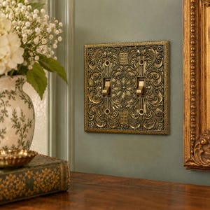 May include: A double toggle light switch plate featuring an intricate, raised antique brass finish with ornate floral and scrollwork patterns. The rectangular plate has a textured border and two metallic toggle switches centered within the design.