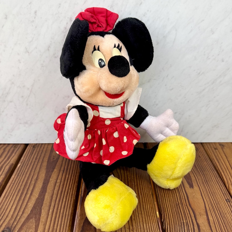 May include: A plush Minnie Mouse doll, featuring black ears, a red bow, and a red dress with white polka dots. The doll has white gloves and yellow shoes. The doll is sitting on a wooden surface.