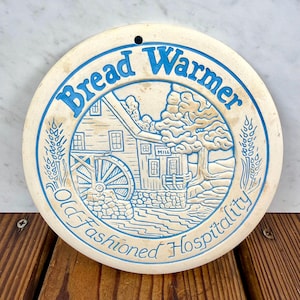 May include: A round, off-white ceramic bread warmer featuring a blue embossed design of a water mill, wheat stalks, and the text "Bread Warmer" and "Old-Fashioned Hospitality". The piece includes a small hole at the top for hanging purposes.