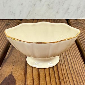 May include: A cream-colored ceramic bowl with a scalloped rim and a gold-painted edge. The bowl features a rounded, fluted body that tapers down to a circular pedestal base, providing a stable foundation for this decorative serving piece.