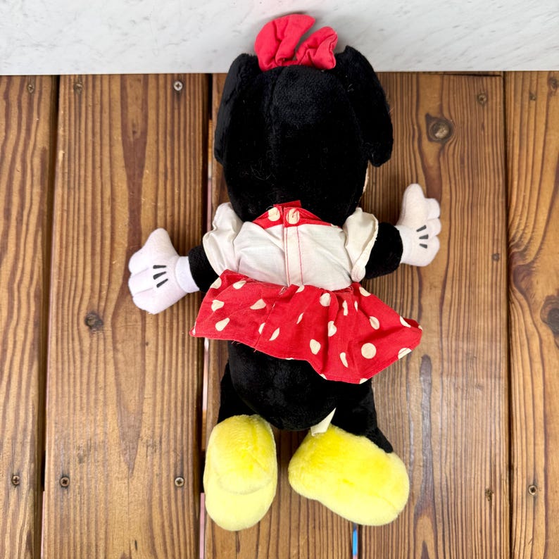 May include: A plush Minnie Mouse doll, viewed from the back, with a black body, red and white polka dot skirt, and a red bow. The doll has white gloves and yellow shoes. The doll is on a wooden surface.