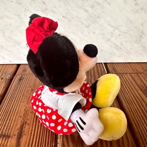 K&ouml;nnte beinhalten: Eine Pl&uuml;schpuppe von Minnie Mouse, mit schwarzem Kopf und Ohren, einer roten Schleife und einem rot-wei&szlig; gepunkteten Kleid. Die Puppe hat wei&szlig;e Handschuhe und gelbe F&uuml;&szlig;e, die auf einer Holzoberfl&auml;che sitzen.