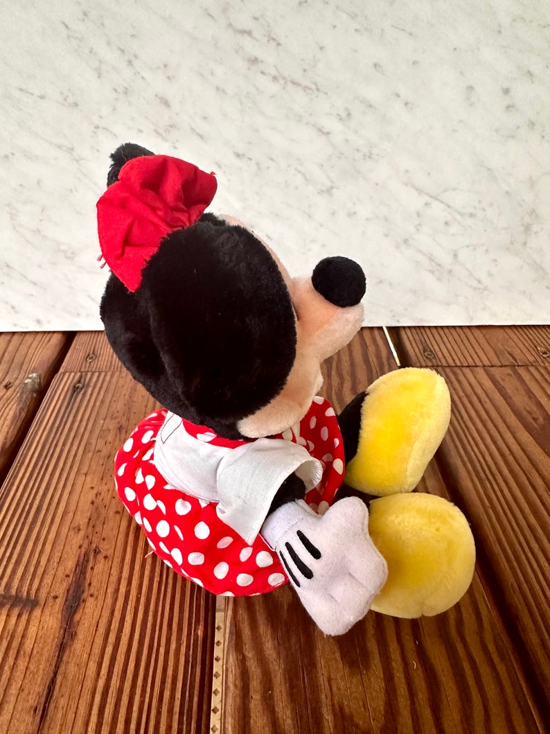 K&ouml;nnte beinhalten: Eine Pl&uuml;schpuppe von Minnie Mouse, mit schwarzem Kopf und Ohren, einer roten Schleife und einem rot-wei&szlig; gepunkteten Kleid. Die Puppe hat wei&szlig;e Handschuhe und gelbe F&uuml;&szlig;e, die auf einer Holzoberfl&auml;che sitzen.