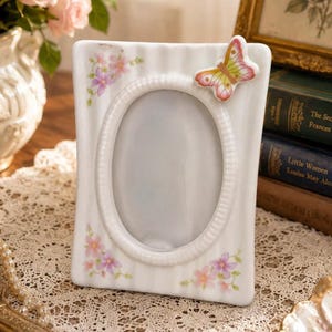 Puede incluir: Un marco de fotos de cerámica blanca con textura acanalada vertical, una abertura central ovalada y un adorno de mariposa en relieve en la esquina superior derecha. El marco está decorado con racimos de flores pintadas en rosa y violeta.