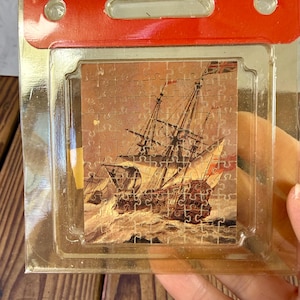 Può includere: Un piccolo puzzle con un'illustrazione vintage di una nave a vela in mare. Il puzzle è in una confezione di plastica trasparente con la parte superiore rossa. La nave è marrone e bianca, con uno sfondo di mare in tempesta.