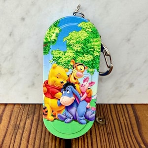 Puede incluir: Un llavero colorido con Winnie the Pooh y sus amigos. El llavero ovalado tiene un fondo de cielo azul y árboles verdes. Los personajes incluyen a Winnie the Pooh, Tigger, Piglet y Eeyore. Un cierre plateado está adjunto en la parte superior.